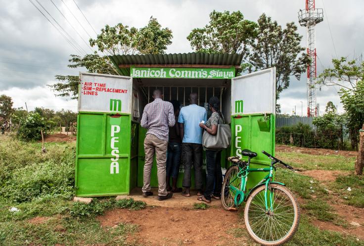 Three people stand at corrugated tin stand advertising mobile services in a field in Kenya