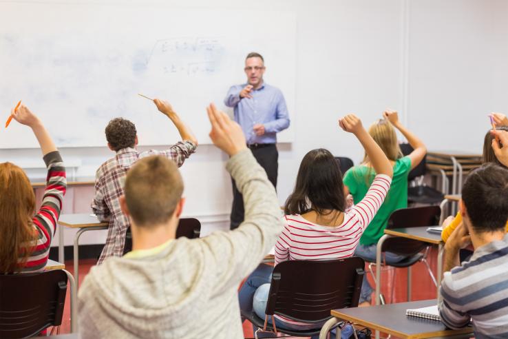 The back of students sitting in desks raising hands, with a teacher at the white board.