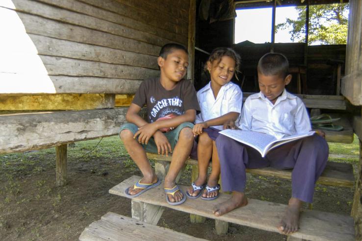 Two boys and a girl shit on the steps of a house reading a large book.