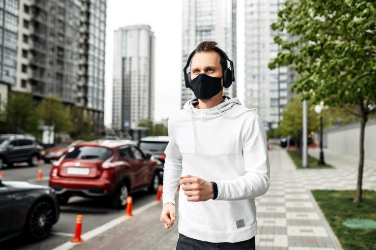 A young man wearing a fit bit, black surgical mask, and white hoodie runs on the sidewalk with cars and buildings in the background.