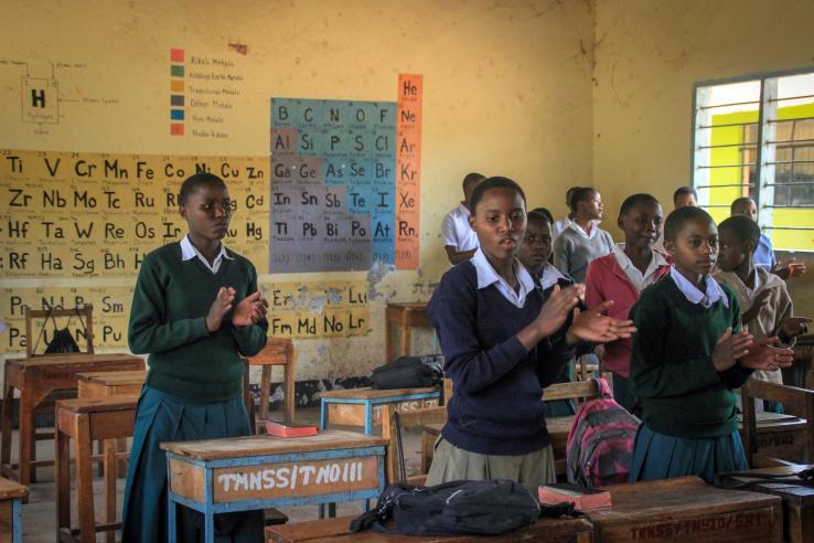 Students stand behind their desks in a classroom clapping their hands together. 