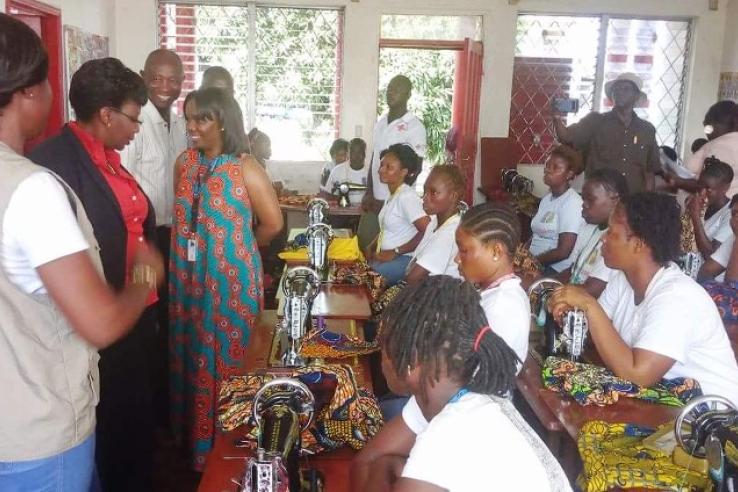 Women in Liberia sit at tables with sewing machines and cloth in front of them. Instructors, including one wearing a vest from the Liberian Red Cross, smile at the front of the room.