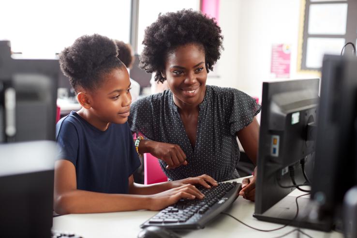 A teenage girl types on a computer while a woman looks on and smiles.
