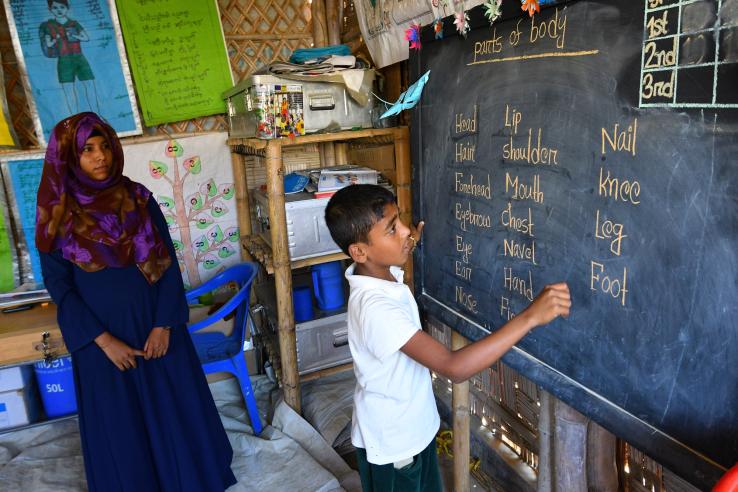 A Bangladeshi child writes on a school blackboard as a teacher watches.