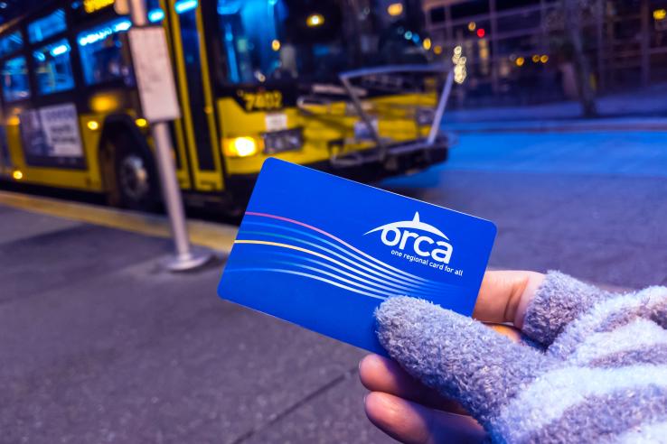 A gloved hand holds a blue ORCA transit card in front of a bus stop where a bus is parked.