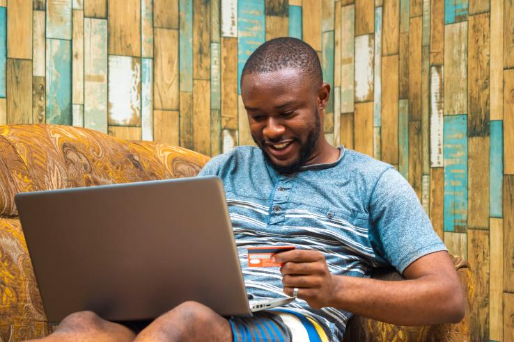 Young black man sitting on a couch and making payments with his card on his laptop