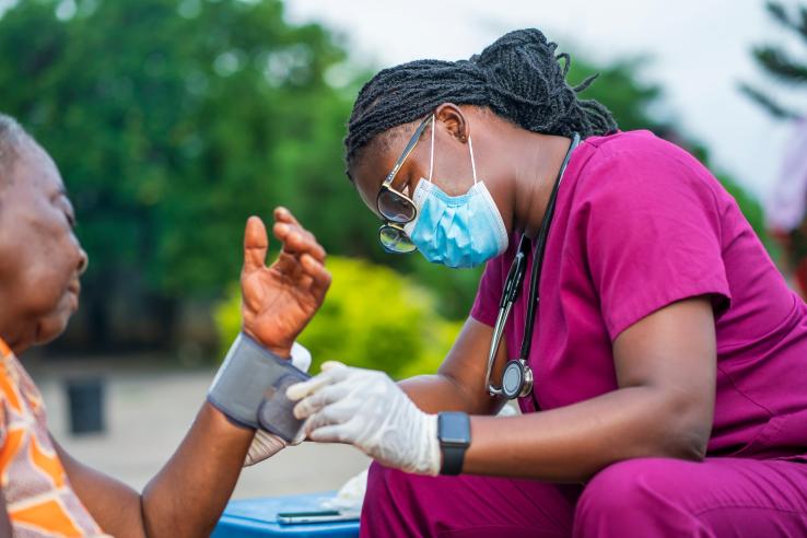 A health worker in Zambia wearing magenta scrubs and blue medical mask checks patient's blood pressure