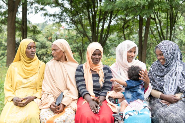 Women sit in a row outside, talking to each other for J-PAL Africa Evaluation Summary