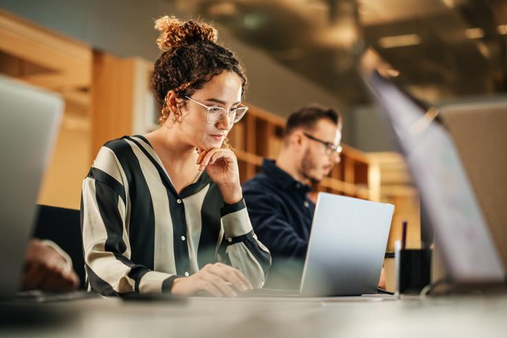Female youth with glasses and curly brown hair looks at her laptop screen with hand to chin in cafe in Denmark