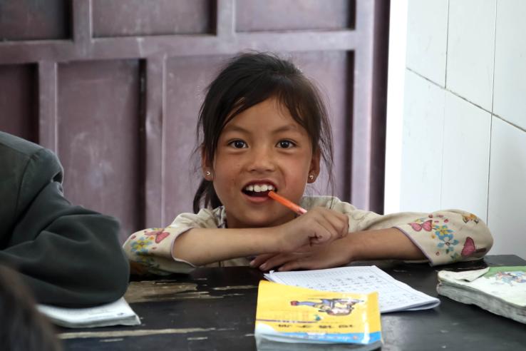 A young Chinese girl sits at desk with pen in mouth and educational workbooks in front of her