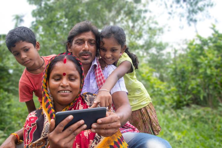A woman sits with her family, showing them something on her smartphone as they gather around her.