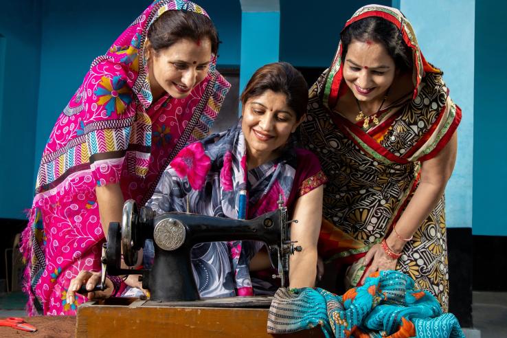 Three women in colorful sarees gather around a sewing machine in India, smiling as one of them works on a piece of fabric.