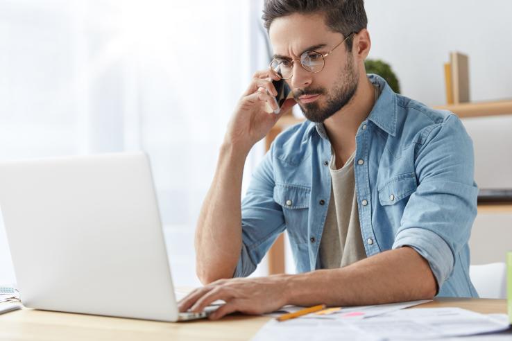 Man in denim shirt sits at a laptop and has a phone up to his ear.