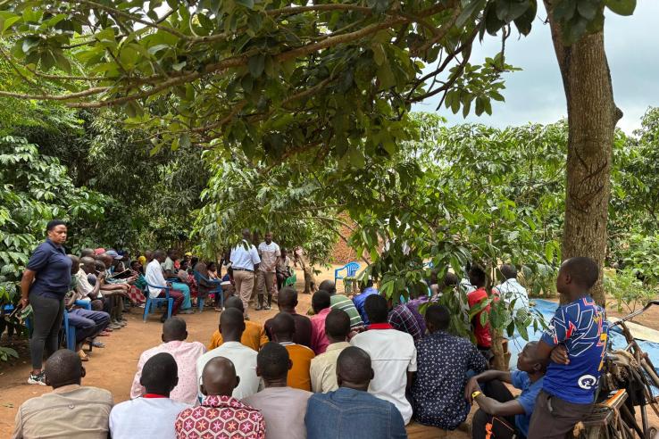 A group of villagers sits in plastic chairs while two men stand and demonstrate agricultural techniques.