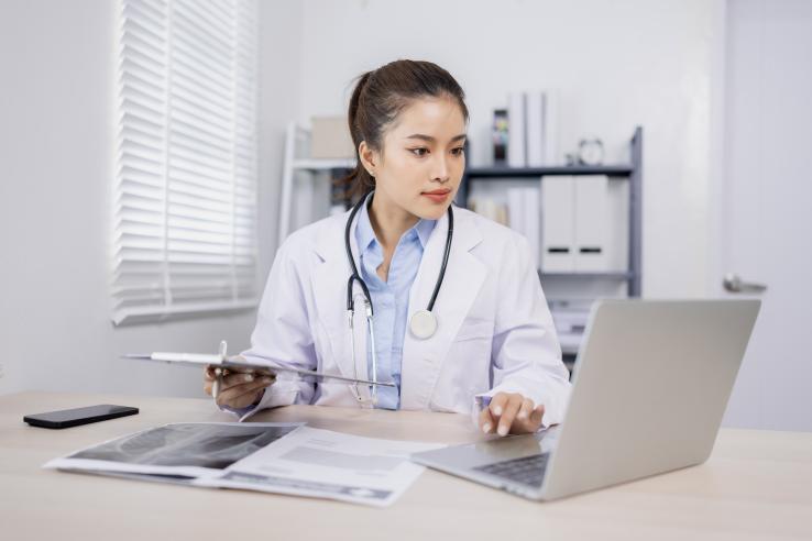 female doctor in white coat sits at desk in front of laptop