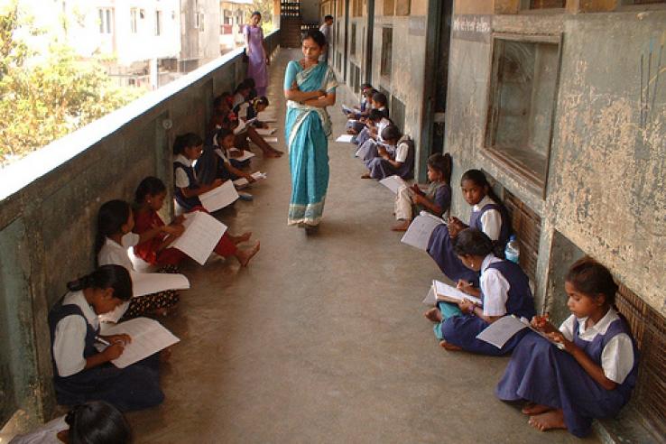 Uniformed students sit on school porch as teacher in sari watches