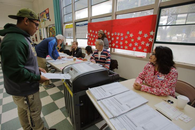 Man feed ballot into ballot box while volunteers help voters in background