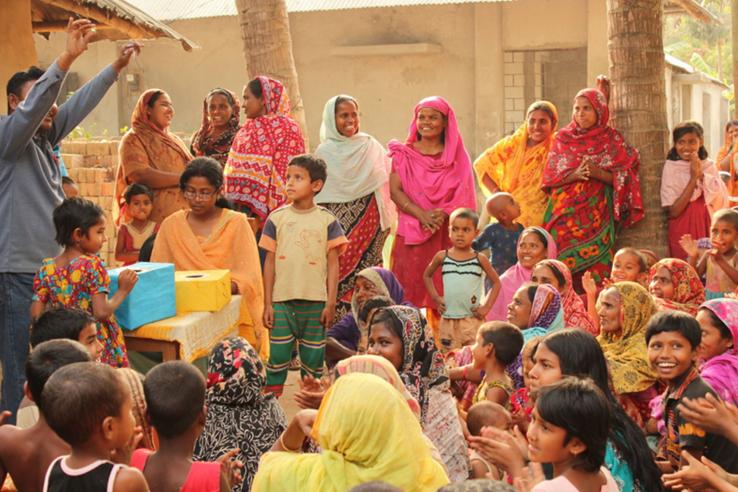 Crowd of women in bright saris watch man hold up token from cardboard box