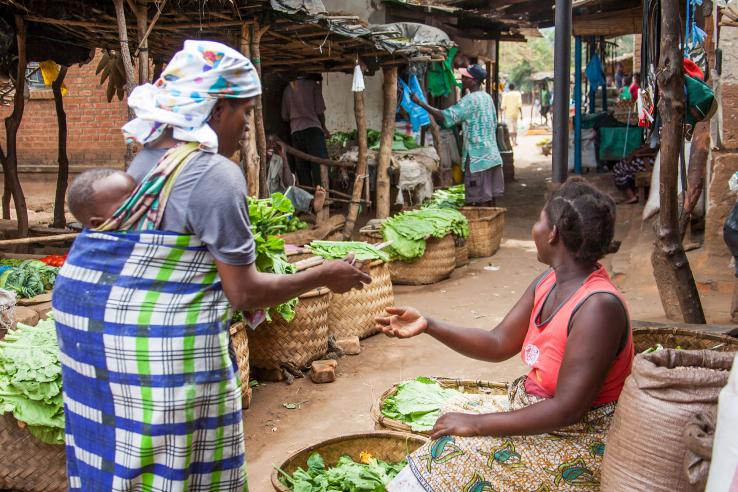 Woman carrying baby on her back pays for her greens at a rural market in Malawi