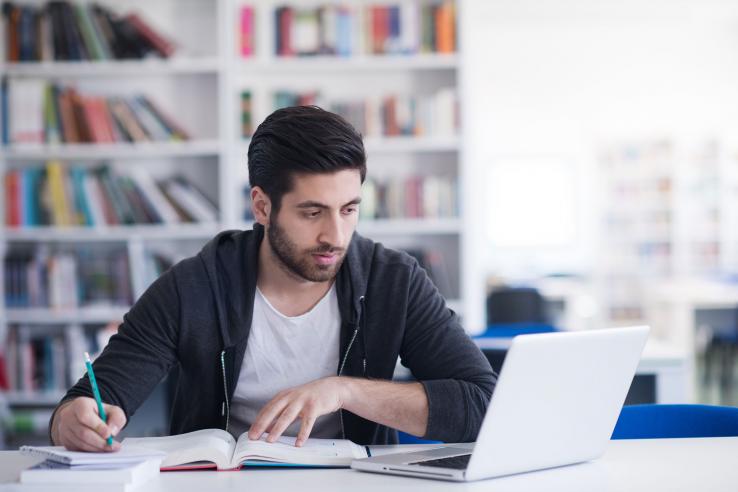 A man sits at a desk, looking at laptop screen while writing notes