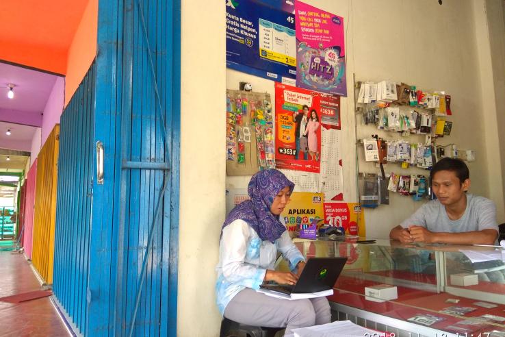 A man stands behind a counter that displays phone cards in a small shop. A woman sits in front of the counter typing on a laptop.