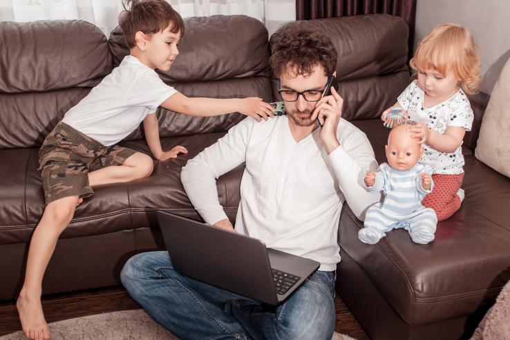 A dad works on his laptop on the couch while children climb all over him.