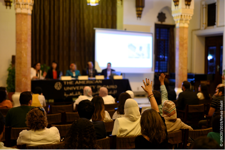 A table of panelists face an audience, several of whom are raising hands