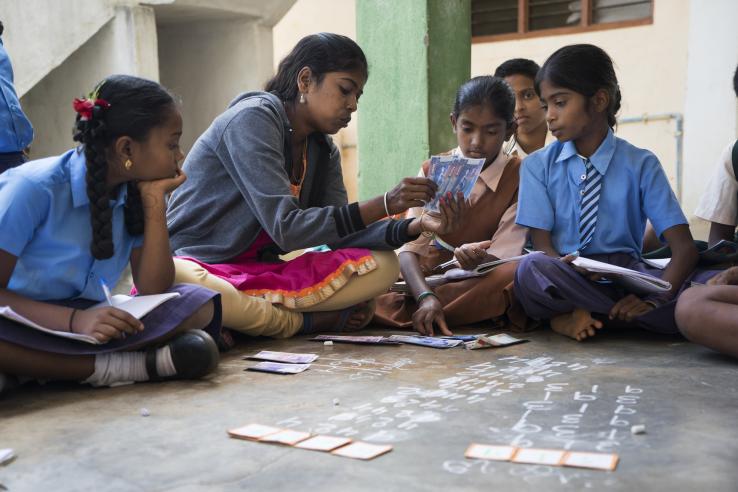 A teacher sits on the ground in a circle of children. She is demonstrating to a student.