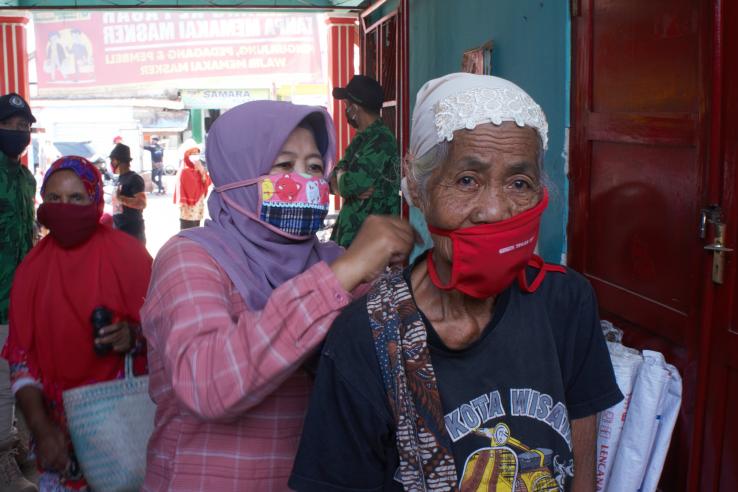 A woman wearing a mask helps an older woman to tie on her mask.