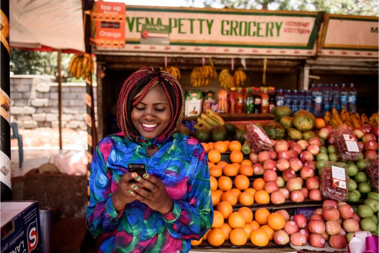 A woman uses a mobile phone to make a cash transfer in front of a fruit stand.