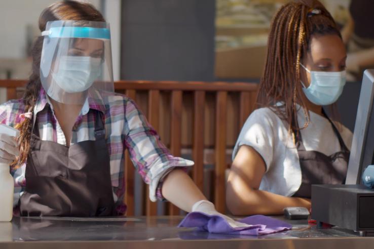 Two female cashiers wearing face masks clean a counter.
