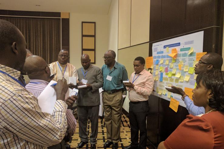 Nine people having a conversation stand around a board covered in post-it notes