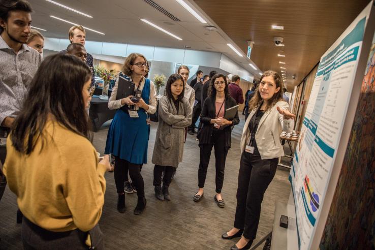 Alejandra Bellating presents a research poster to an audience standing around her.