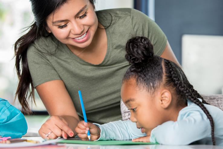 A women sits at a desk with a little girl as she writes something down with a pencil
