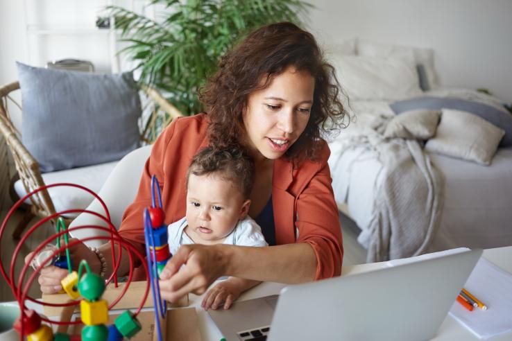 A woman at a desk with a baby on her lap looks at her laptop screen while the baby plays