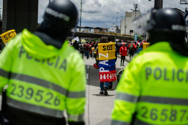 Two police officers in riot gear look at a protester wearing shield that says "Paz"