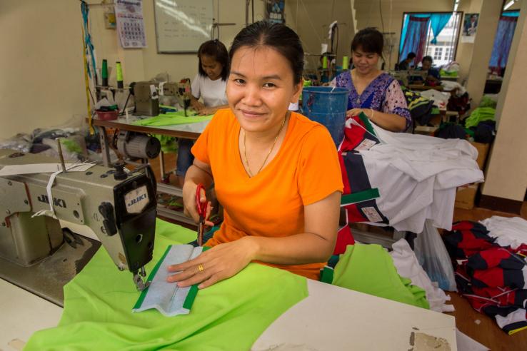 A woman sitting at a sewing desk smiles at the camera while cutting cloth with scissors