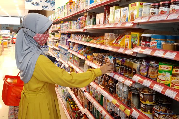 A woman wearing a face mask pulls a food item off of the shelf in a grocery store.