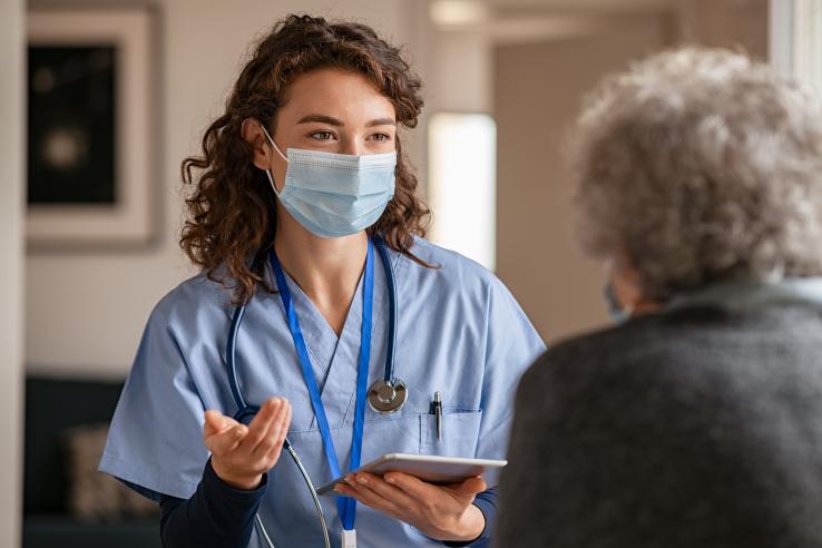 A woman wearing scrubs and a mask speaks to an elderly person