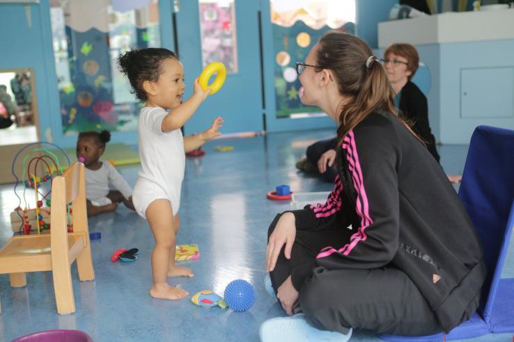 A young child in a daycare setting shows an adult caretaker a yellow plastic ring toy.