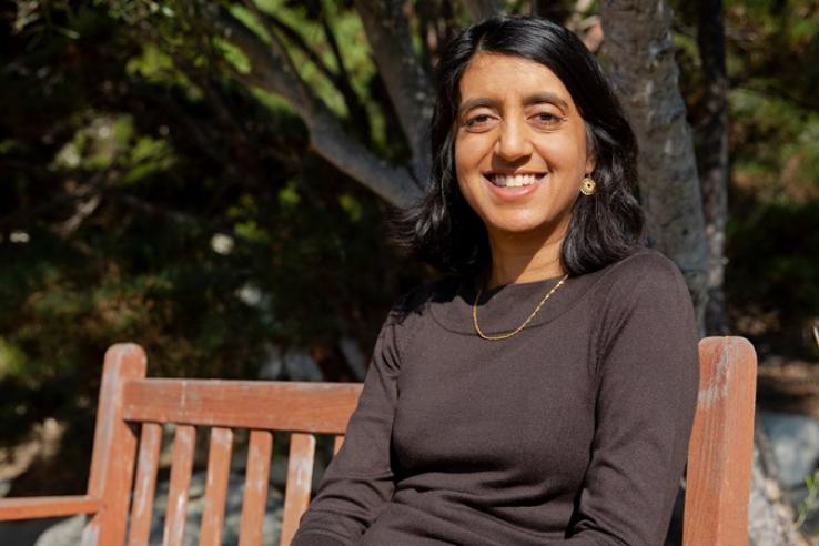 Seema Jayachandrani sits on a wooden bench outside and smiles at the camera.