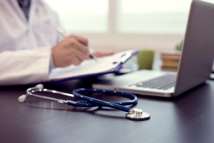 A doctor sits at a computer looking at a chart on a clipboard