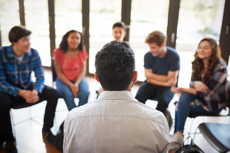 A circle of people sitting in chairs face a man sitting on a chair in front of them