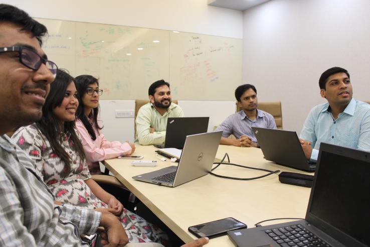 A group of six people sit around a table with laptops, looking forward