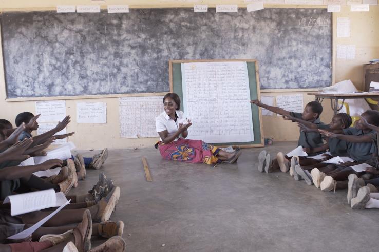 A teacher sits on the floor by a chalk board. Students on either side of her raise their hands.