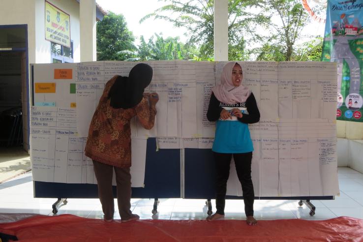 Two women wearing head scarves stand in front of a white board at the front of a classroom. 