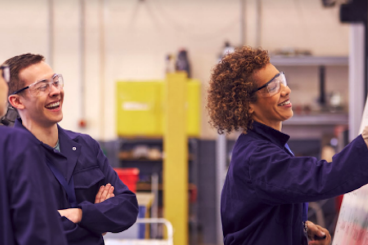 Adult workers wearing blue coveralls gather around a whiteboard in an industrial setting.