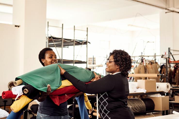 Two smiling women exchange a roll of fabric inside a textile factory in Zimbabwe.