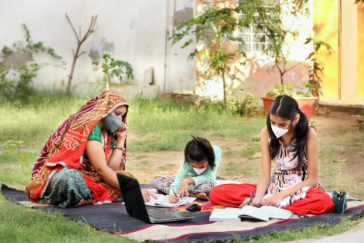 A woman sits with children outdoors on a mat with a laptop and books. Everyone is wearing a mask.