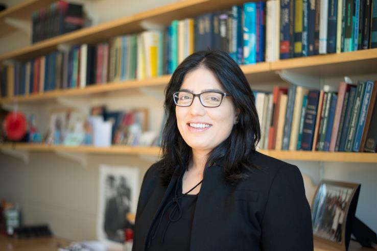 A woman stands in front of a bookcase wearing a black jacket and glasses. 